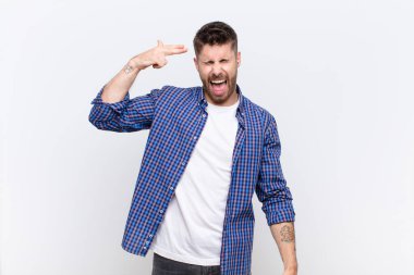 Genç adam mutsuz ve stresli görünüyor, intihar jesti elle silah işareti yapıyor, düz renkli duvara işaret ediyor.