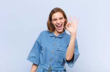 Genç sarışın kadın kendini başarılı ve tatmin olmuş hissediyor, ağzı açık gülümsüyor, düz renkli duvara karşı elle işaret veriyor.