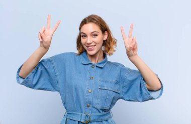 Genç sarışın kadın gülümsüyor ve mutlu görünüyor, arkadaş canlısı ve memnun, iki elini de düz duvara dayayıp zafer ya da barış işareti yapıyor.