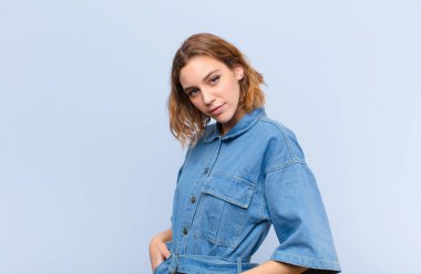 Genç sarışın kadın gururlu, kendinden emin, havalı, küstah ve küstah görünüyor, gülümsüyor, düz renkli duvara karşı başarılı hissediyor.