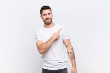 Genç adam neşeyle gülümsüyor, mutlu hissediyor ve yan ve yukarı doğru işaret ediyor, düz renkli duvara karşı fotokopi uzayındaki nesneleri gösteriyor.