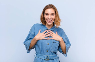 Genç sarışın kadın mutlu, şaşırmış, gururlu ve heyecanlı görünüyor. Düz renkli duvara işaret ediyor.