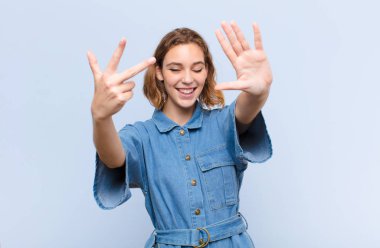 Genç sarışın kadın gülümsüyor ve arkadaş canlısı görünüyor. Sekiz ya da sekiz numarayı gösteriyor. Elleri önde, düz renkli duvara doğru sayıyor.