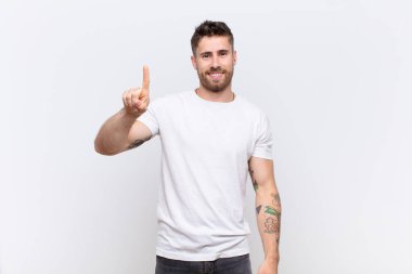 Genç adam gülümsüyor ve arkadaş canlısı görünüyor, bir numara ya da ilk eli önde, düz renkli duvara doğru sayıyor.