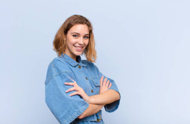 Çapraz kollarıyla kameraya gülümseyen genç sarışın kadın ve mutlu, kendinden emin, tatmin olmuş bir ifade, düz renkli duvara bakan yanal bir bakış.