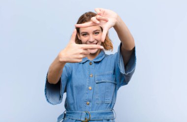 Genç sarışın kadın mutlu, arkadaş canlısı ve pozitif hissediyor, gülümsüyor ve düz renkli duvara karşı elleri ve fotoğraf çerçevesi yapıyor.