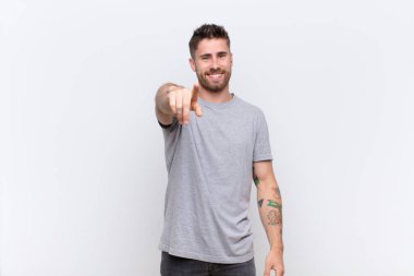 Genç adam kameraya tatmin olmuş, kendine güvenen, dost canlısı bir gülümsemeyle bakıyor, düz renkli duvara karşı seni seçiyor.