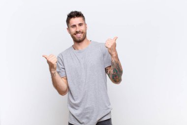 Genç adam neşeyle gülümsüyor ve mutlu görünüyor, kaygısız ve pozitif hissediyor iki başparmağı düz renkli duvarda