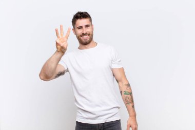 Genç adam gülümsüyor ve arkadaş canlısı görünüyor, üç ya da üç numarayı gösteriyor, düz renkli duvara doğru sayıyor.