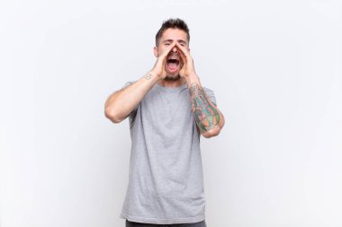Genç handosme adam mutlu, heyecanlı ve pozitif hissediyor, elleri ağızının yanında büyük bir haykırış, düz renkli duvara karşı bağırıyor