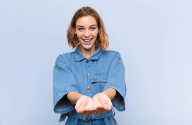 Genç sarışın kadın mutlu bir şekilde gülümsüyor, arkadaş canlısı, kendinden emin, pozitif bakışlı, düz renkli duvara karşı bir nesne veya konsept sunuyor ve gösteriyor.