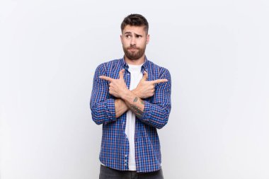 Genç adam şaşkın ve şaşkın görünüyor, güvensiz ve zıt yönleri işaret ediyor düz renk duvarına karşı şüpheyle