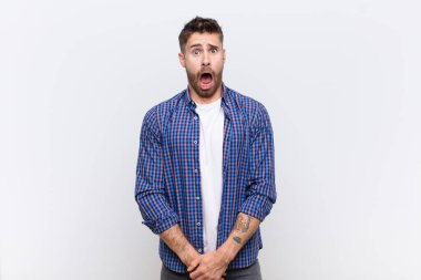 Genç handosme adam korkmuş ve şok olmuş hissediyor, ağzı açık, düz renkli duvara karşı şaşkınlık içinde.