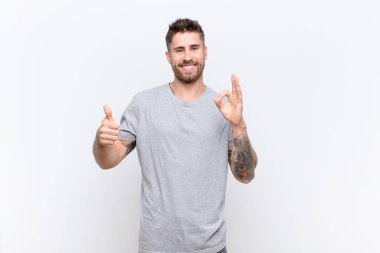 Genç adam kendini mutlu, şaşkın, tatmin olmuş ve şaşırmış hissediyor, tamam gösteriyor ve jestleri parmakla gösteriyor, düz renkli duvara gülümsüyor.