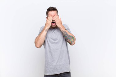 Genç adam gülümsüyor ve mutlu hissediyor, iki eliyle gözlerini kapatıyor ve düz renkli duvara karşı inanılmaz bir sürpriz bekliyor.