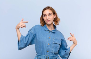 Genç sarışın kadın gururlu, kibirli, mutlu, şaşırmış ve tatmin olmuş görünüyor, kendini gösteriyor, düz renkli duvara karşı kazanan gibi hissediyor.