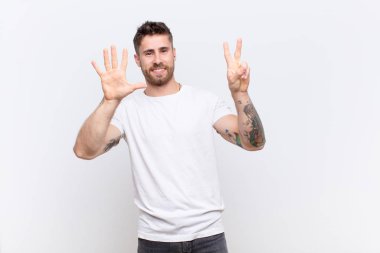 Genç adam gülümsüyor ve dost canlısı görünüyor, 7 ya da 7 numarayı gösteriyor, eli önde, düz renkli duvara doğru geri sayıyor.