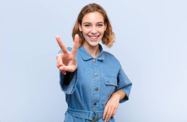 Genç sarışın kadın gülümsüyor ve mutlu görünüyor, kaygısız ve pozitif, bir eliyle düz duvara karşı zafer ya da barış işareti yapıyor.