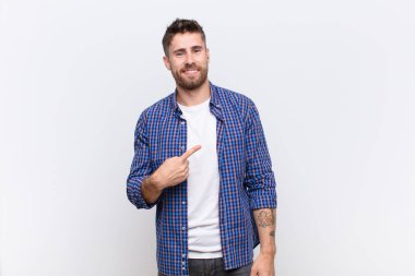 Genç adam gururlu, kendinden emin ve mutlu görünüyor, gülümsüyor ve kendini işaret ediyor ya da düz renk duvara karşı bir numara işareti yapıyor.