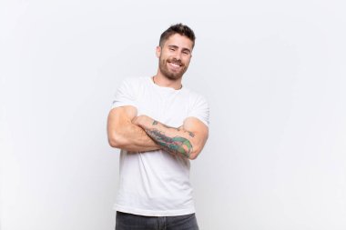 Genç adam kollarını kavuşturup mutlu bir şekilde gülüyor, rahat, pozitif ve tatmin olmuş bir pozla düz renkli duvara karşı.