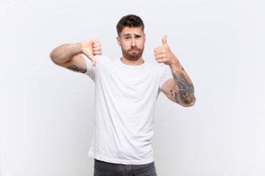 Genç adam kafası karışmış, bilgisiz ve kararsız hissediyor, iyi ve kötü seçenekleri farklı seçeneklerde tartıyor veya düz renkli duvarlara karşı seçenekleri tartıyor.