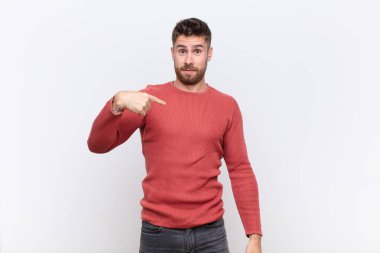 Kafası karışmış, kafası karışmış ve kendine güveni olmayan genç bir adam kendini merak edip bana mı soruyor? yassı renk duvarına karşı