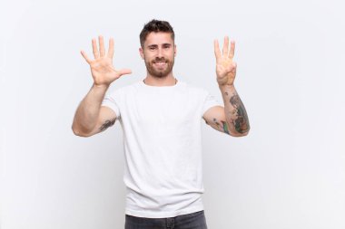 Genç adam gülümsüyor ve arkadaş canlısı görünüyor. Sekiz ya da sekiz numarayı gösteriyor. Elleri önde, düz duvara doğru sayıyor.