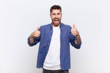 Genç adam gülümsüyor mutlu, pozitif, kendine güvenen ve başarılı, iki başparmağı düz renkli duvara dayalı.