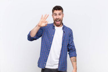 Genç adam kendini başarılı ve tatmin olmuş hissediyor, ağzı açık gülümsüyor, düz renkli duvara karşı elle işaret veriyor.