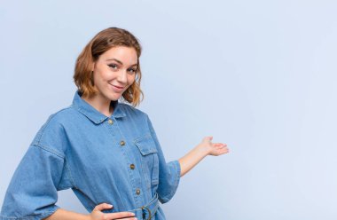 Genç sarışın kadın kendini mutlu ve neşeli hissediyor, gülümsüyor ve seni hoş karşılıyor, düz renkli duvara karşı dostça bir jestle içeri davet ediyor.