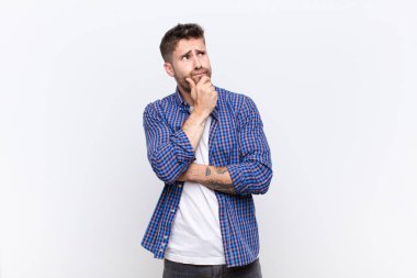 Genç adam düşünüyor, kuşkulu ve şaşkın hissediyor, farklı seçeneklerle, düz renkli duvara karşı hangi kararı vereceğimi merak ediyor.