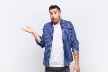 Genç handosme adam şaşırmış ve şok olmuş görünüyor, çene düşmüş düz renkli duvara karşı bir el açık bir nesne tutuyor