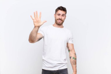 Genç adam gülümsüyor ve arkadaş canlısı görünüyor, 5 ya da 5 numarayı gösteriyor, düz renkli duvara doğru sayıyor.