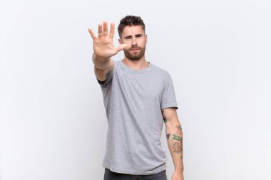 Genç adam ciddi, sert, hoşnutsuz ve kızgın görünüyor. Avuç içi açık, düz renkli duvara karşı jest yapıyor.