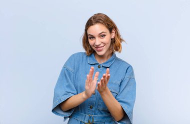 Genç sarışın kadın mutlu ve başarılı hissediyor, gülümsüyor ve alkışlıyor, düz renkli duvara karşı alkış ile tebrik ediyor.