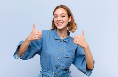 Genç sarışın kadın neşeyle gülümsüyor ve mutlu görünüyor, kaygısız ve pozitif hissediyor iki başparmağı düz renkli duvarda.