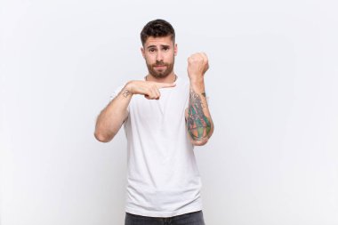 Genç adam sabırsız ve kızgın görünüyor, saati gösteriyor, dakiklik istiyor, düz renkli duvara karşı zamanında olmak istiyor.