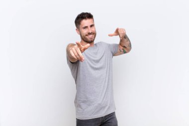 Genç adam kendini mutlu ve kendinden emin hissediyor, kamerayı iki eliyle gösteriyor ve gülüyor, düz renkli duvara karşı seni seçiyor.