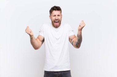 Genç adam son derece mutlu ve şaşırmış görünüyor, başarıyı kutluyor, bağırıyor ve düz renkli duvara zıplıyor.