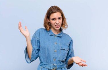 Genç sarışın kadın şaşkın, şaşkın ve stresli görünüyor, farklı seçenekler arasında merak ediyor, düz renk duvarına karşı kararsız.