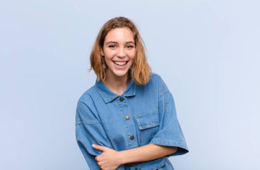 Genç sarışın kadın utangaç ve neşeli bir şekilde gülüyor, arkadaş canlısı ve olumlu ama düz renkli duvara karşı güvensiz bir tutum sergiliyor.