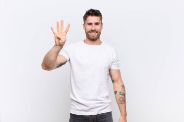 Genç adam gülümsüyor ve arkadaş canlısı görünüyor, dört ya da dört numarayı gösteriyor, düz renkli duvara doğru sayıyor.