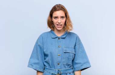 Genç sarışın kadın şok olmuş, kızgın, sinirli ya da hayal kırıklığına uğramış, ağzı açık ve düz renkli duvara karşı öfkeli görünüyor.