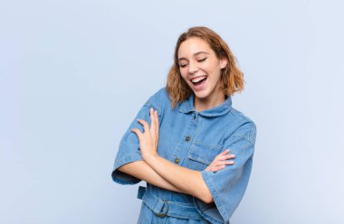 Sarışın genç bir kadın kollarını kavuşturup mutlu bir şekilde gülüyor, rahat, pozitif ve tatmin olmuş bir pozla düz renkli duvara yaslanıyor.