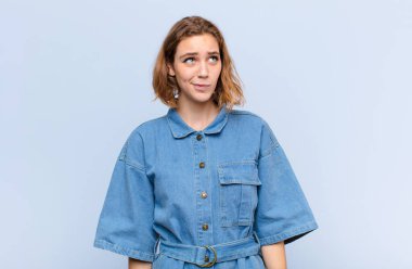 Genç sarışın kadın şaşkın ve şaşkın görünüyor, merak ediyor ya da bir sorunu çözmeye çalışıyor ya da düz renkli duvara karşı düşünüyor.