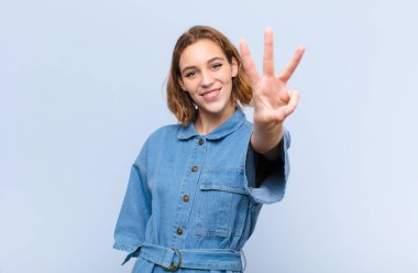 Genç sarışın kadın gülümsüyor ve arkadaş canlısı görünüyor. Üç ya da üç numarayı gösteriyor. Elleri önde, düz renkli duvara doğru sayıyor.