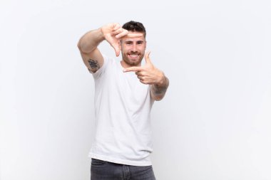 Genç adam mutlu, arkadaş canlısı ve pozitif hissediyor, gülümsüyor ve düz renkli duvara karşı elleriyle bir portre veya fotoğraf çerçevesi yapıyor.