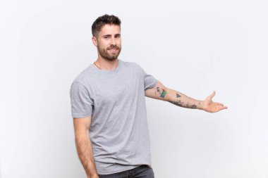 Genç adam kendini mutlu ve neşeli hissediyor, gülümsüyor ve seni selamlıyor, düz renkli duvara karşı dostça bir jestle içeri davet ediyor.