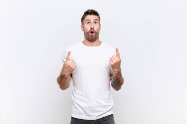 Genç adam şaşırmış, şaşırmış ve ağzı açık görünüyor, iki eliyle yukarıya bakıyor düz renkli duvara uzayı kopyalamak için.