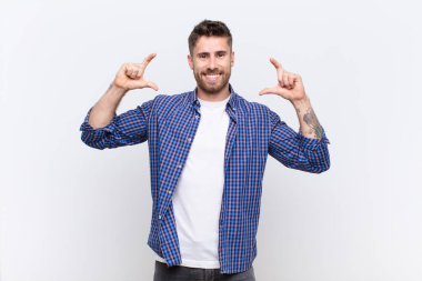 Genç adam kendi gülümsemesini çerçeveletiyor ya da özetliyor, iki eliyle, pozitif ve mutlu görünüyor, düz renkli duvara karşı sağIık kavramı